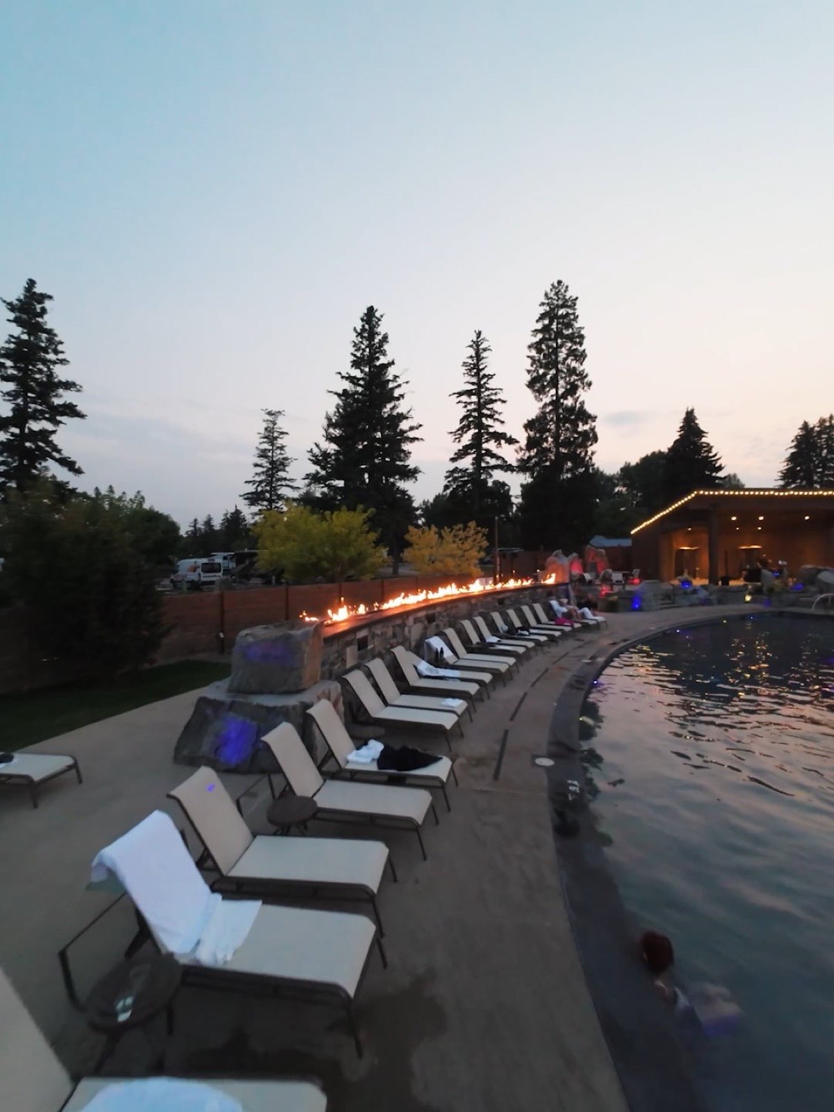 White loungers lined along the lit outdoor pool at Bozeman Hot Springs at dusk, pines behind