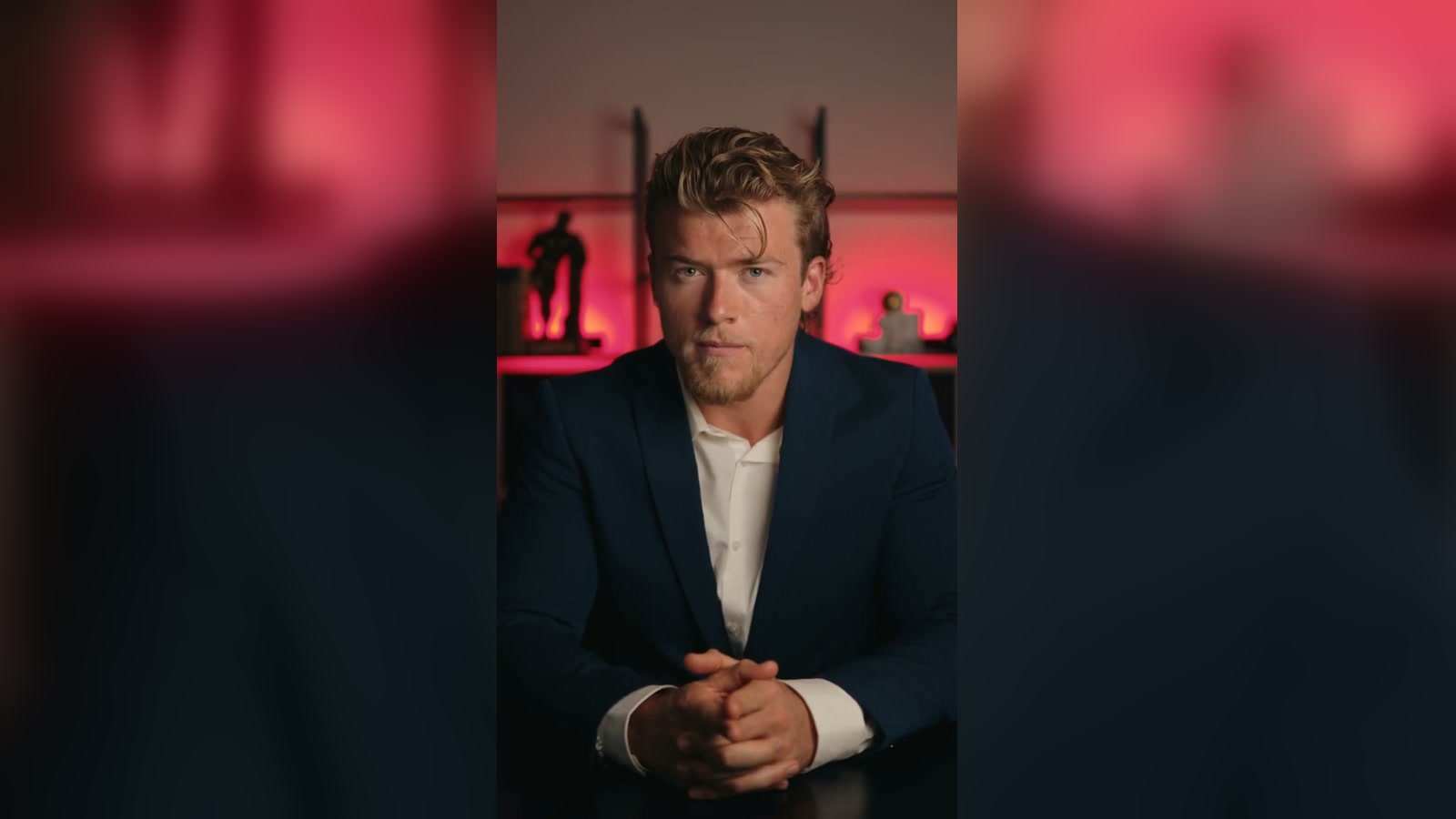 Brandon Clark in a navy blazer, hands clasped, seated in front of a red-lit shelf unit