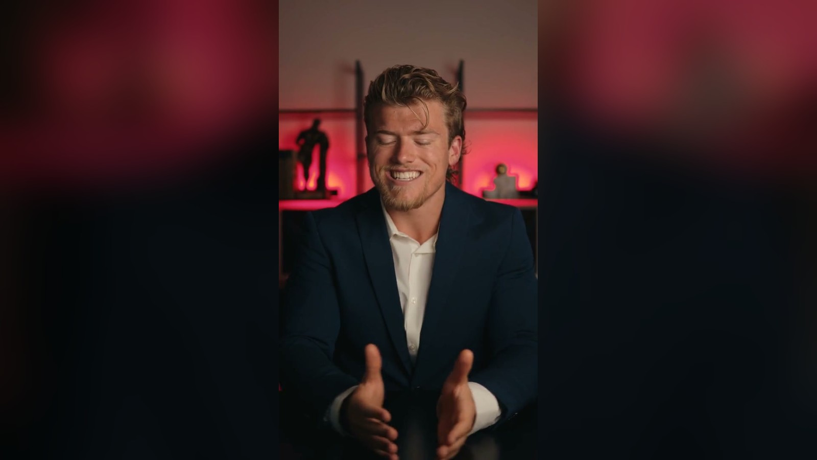 Brandon Clark in a navy blazer laughing mid-take in front of a red-lit shelf unit