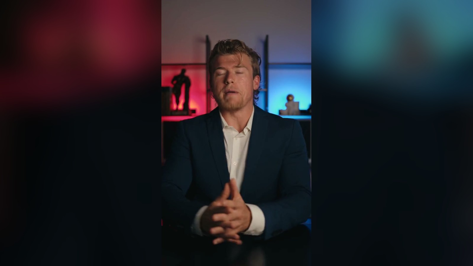 Brandon Clark in a navy blazer talking to camera in front of a blue and red lit shelf unit