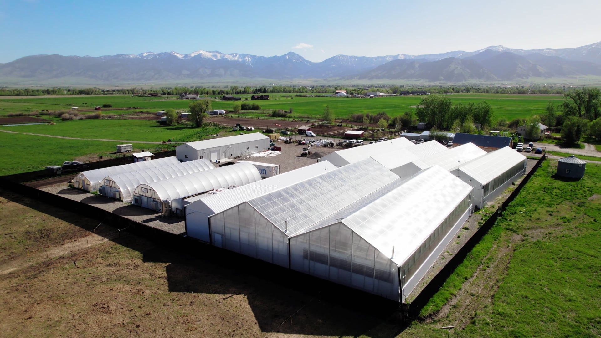 Aerial over Collective Elevation's white greenhouses on a green field with the Bridger range behind